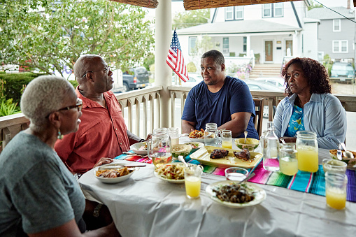 Casually dressed family members in 40s, 50s, and 60s sitting at dining table on veranda of Rockaway Beach home and pausing from eating to converse.