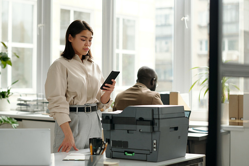 Young serious businesswoman looking at smartphone screen in office while standing by workplace with xerox machine and texting