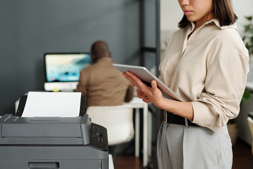 Focus on tablet held by young elegant businesswoman computing in office while standing by xerox machine against African American male coworker