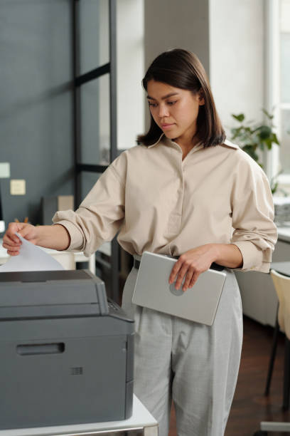 Young brunette businesswoman with tablet in hand standing in front of copier stock photo