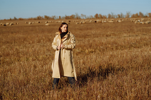 Young woman in a coat standing in a field among dry grass, enjoying nature. Urban style and street fashion. Concept of nature, fashion, freedom. Lifestyle.