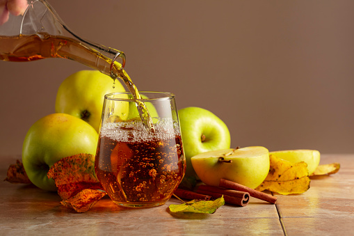 Apple juice is poured from a bottle into a glass. On a ceramic table ripe apples, cinnamon sticks, and dried-up leaves. Beige background with copy space.