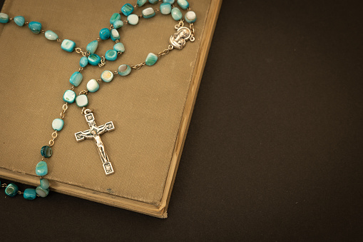 Rosary with blue prayer beads on a tan book all on a black background with copy space