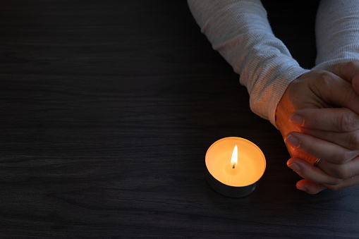 Hands of a woman folded in prayer beside a single tea light candle on a dark wood table with copy space