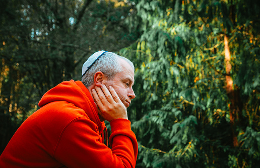 Color image depicting a Jewish man wearing a yarmulke, or kippah, while meditating and lost in thought in the park. The young man is dressed casually in red hoodie, blue denim jeans and black gilet.