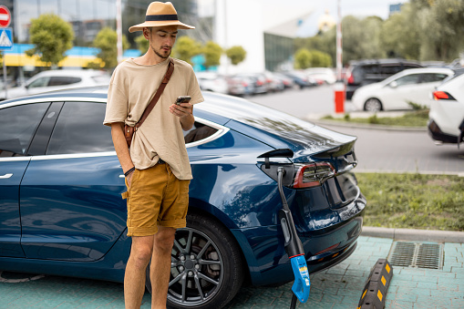Man waits for his car to be charged, while standing with phone on a public charging station outdoors. Concept of travel by electric car and green energy for driving