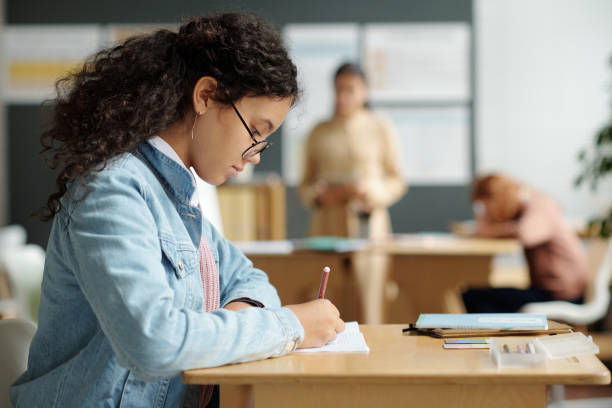 istock Side view of serious schoolgirl in eyeglasses making notes in copybook 1741111053