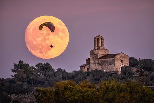 candid photo of a paramotor flying in front of the full moon near a Romanic church at sunset in Catalonia, Spain