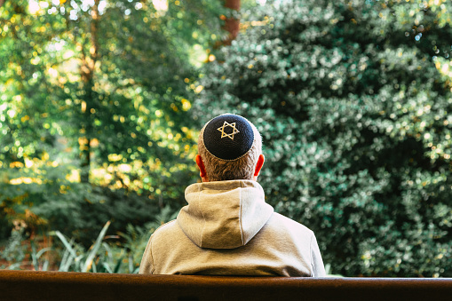 Color image depicting a Jewish man wearing a yarmulke, or kippah, while meditating and lost in thought in the park. The young man is dressed casually in grey hoodie and blue denim jeans.