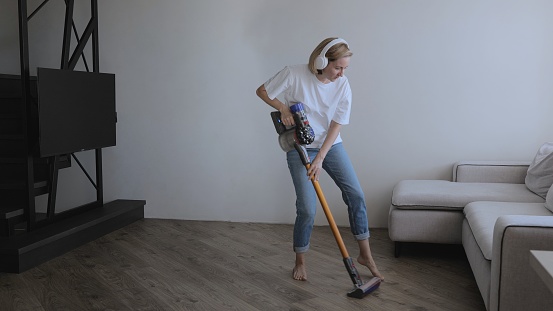 A young woman is cleaning and dancing with a vacuum cleaner in her apartment. Energetic woman doing housework, cleaning the living room. Cheerful woman listens to music while doing housework