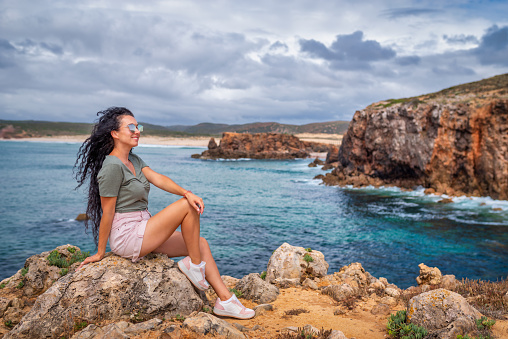 Beautiful woman seated over the rocks in front of Bordeira beach at Pontal da Carrapateira in a cloudy beach, Algarve, Costa Vicentina, Portugal