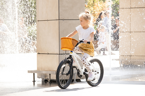 Cute little caucasian blonde girl riding a white bike with wicker basket in the amusement park. Water drops of a fountain during sunny summer day in the background.