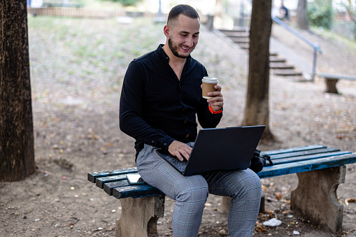 Embracing the outdoors, a young businessman engages in his work on a laptop while savoring coffee in a peaceful public park