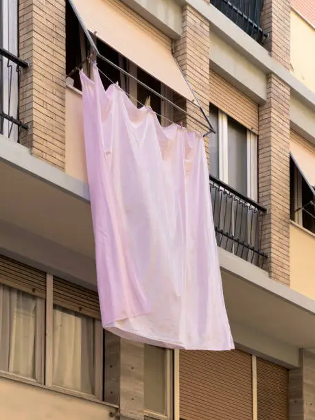 Pink sheet billowing in the wind as it hangs from a clothesline on a balcony. The backdrop of a brick apartment building with shutters encapsulates the essence of Mediterranean lifestyle and culture