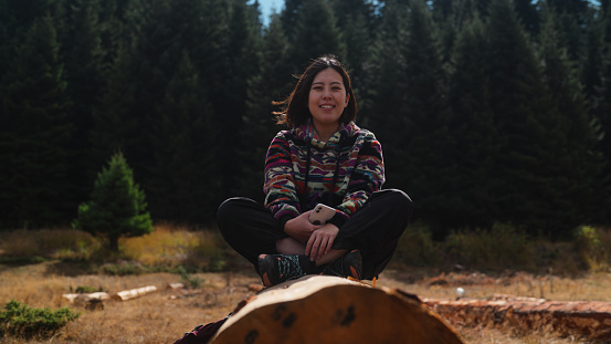 A portrait of a cheerful Asian female solo traveler and hiker is sitting on a tree log and looking at the camera and smiling in nature in mountain.