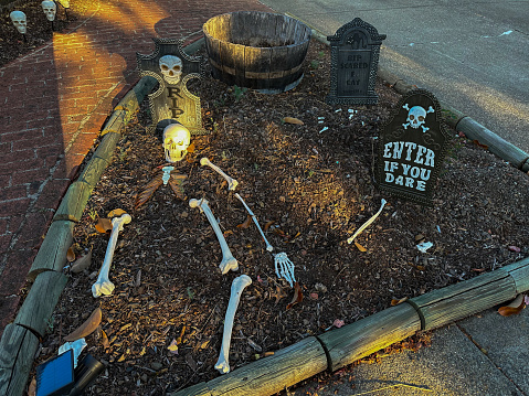 A Halloween display of a skeleton and skull are on a dirt area. Some of the skeleton is buried.  Another skeleton is on the right with a sign saying Enter if You Dare. A headstone is in the back.