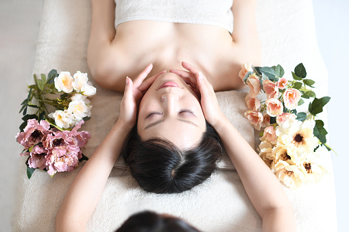 A esthetician working on a young Asian woman lying on her back on a bed receiving a facial massage at a spa