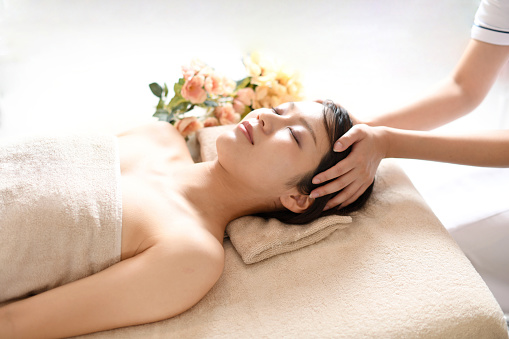 An esthetician working on a young Asian woman lying on her back on a bed receiving a head massage at a spa