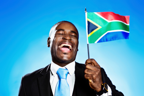 This patriotic, handsome  African businessman or politician laughs and shouts triumphantly as he waves the South African flag against a blue sky. Freedom at last!