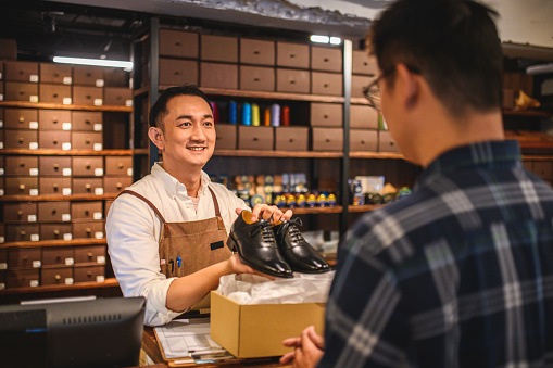 Mid adult Chinese salesman working and selling elegant leather shoes in a designer's boutique at the shopping mall. He is wearing a white shirt and a light brown apron. The young male customer is defocused in the foreground.