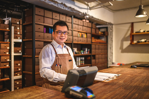 Portrait of a Japanese male business owner working late hours behind the counter. He is indoors in a luxury menswear and accessories store. He is wearing glasses, a light color shirt and a brown apron. He is happy to work hard in order to achieve his goals.