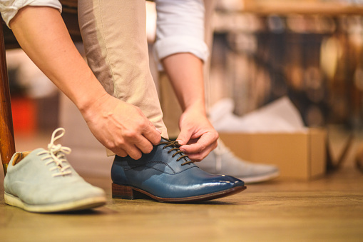 Close-up shot of an asian man trying on natural blue leather shoes. He is tying the shoelaces with his hands. He is indoors in a menswear clothing store at the shopping mall.