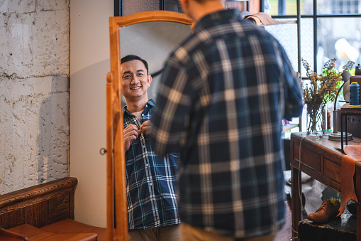 Mid adult asian man trying on a plaid blue shirt in a fitting room of a menswear clothing store. He is buying new clothes to go out on a date. He is looking at himself in the mirror and feeling handsome.