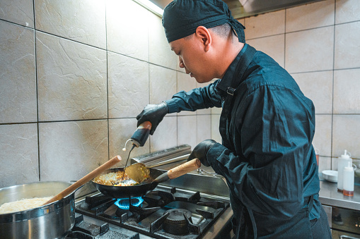 Male chef wearing a black uniform while cooking a delicious lunch. He is creating a fusion of noodles and vegetables that promises to be both fresh and delicious.