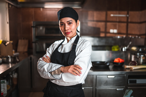 Indoor portrait of a female chef standing in the kitchen. She is wearing a white uniform and has her arms crossed.