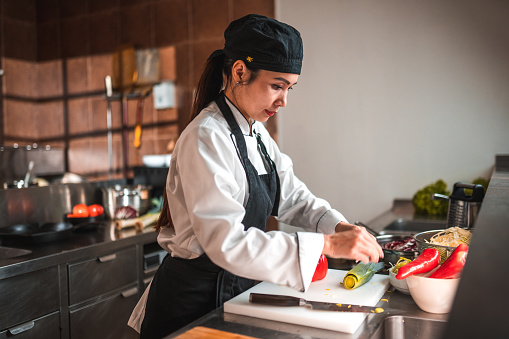 Waist up shot of an asian woman using a knife and chopping a red pepper with quick, precise cuts. She is standing in by the counter and wearing a white uniform.