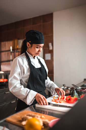 Mature Asian female chef works in the kitchen, slicing a red pepper into small pieces for a dish. She is standing close to the sink and countertop.