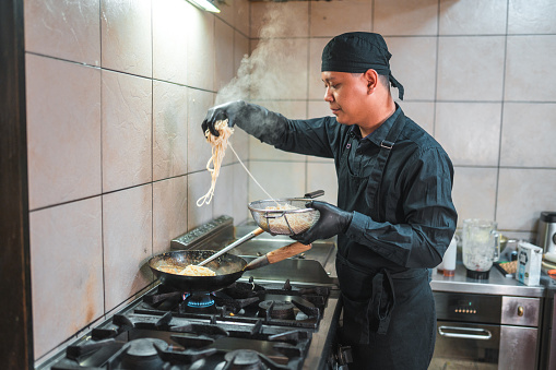 Young chinese chef wearing a black uniform and cooking freshly made noodles in the wok. He is carefully adding them into the kitchen appliance.