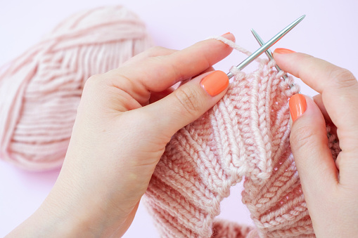 Female hands knit a hat with knitting needles on a pink background close up.