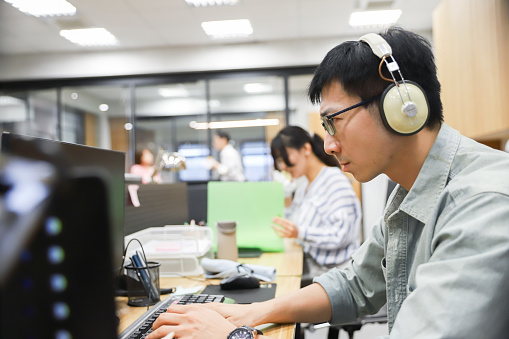 An Asian man wears wired headphones and listens to music while working,There are many colleagues around him, and everyone is busy with their own work.
