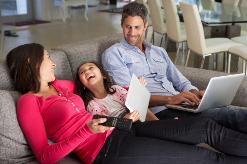 Family On Sofa With Laptop And Digital Tablet Watching TV Smiling And Laughing
