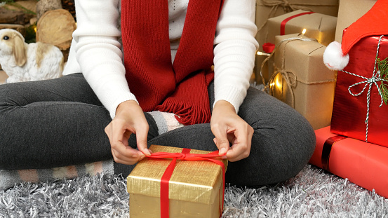 Close up woman hands wrap present gift in cozy living room. Woman wrapping christmas gift box under xmas tree. Asian woman decor bauble ball christmas tree in xmas holiday winter seasonal celebration