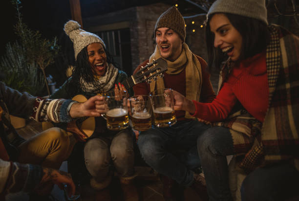 Group of multi-ethnic friends sit around a roaring campfire on a winter evening. They are each dressed in warm clothes , smile and playing the guitar.