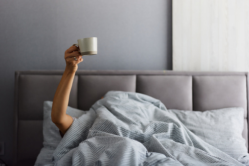 Unrecognizable woman lying covered with duvet in a bed while holding coffee cup high up in her hand. Copy space.