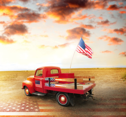 Red vintage pick up truck with American flag in wide open country side with dramatic sunset cloudscape and US flag on ground