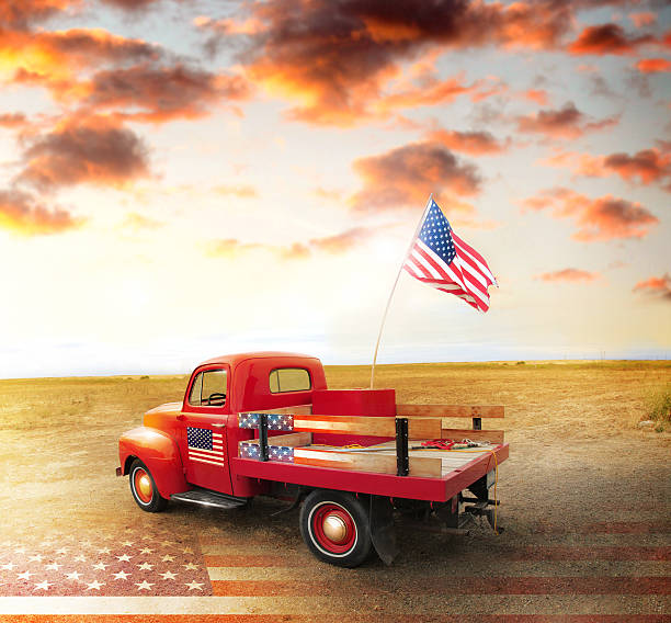 American spirit Red vintage pick up truck with American flag in wide open country side with dramatic sunset cloudscape and US flag on ground vintage american flag stock pictures, royalty-free photos & images