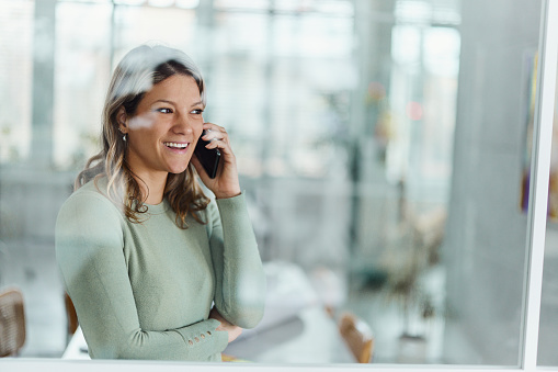 Happy female entrepreneur communicating with someone over smart phone in the office. The view is through glass.