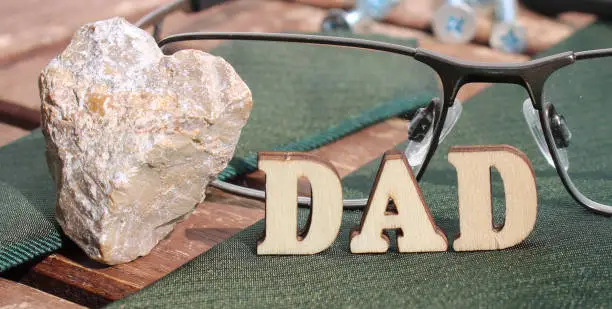 nature wooden letters in the sun on a sunny day. create text DAD from wooden letter alphabet. background is made from naturally wood table, screws, glasses, green tie, stoneheart, tools