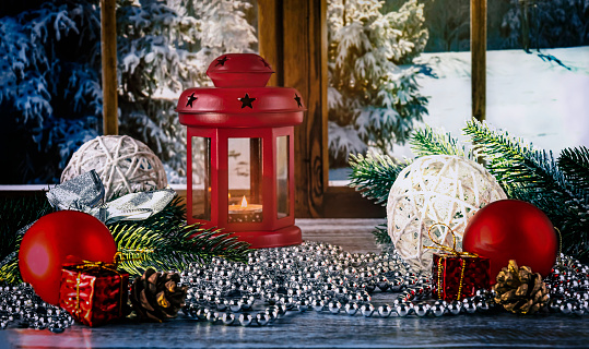 A red lantern with a candle on the background of the window. Christmas red and white balls, necklace, toys and fir branches and cones as decoration. Merry Christmas concept