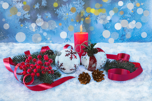 A red candle, Christmas tree toys, Christmas tree branches, rowan, cones on a snowy window background. Festive Christmas composition with red ribbon and bokeh.