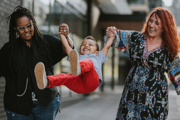 Happy family walking on city sidewalk A multiracial lesbian couple smile as they playfully swing their toddler son in the air as they walk down a city sidewalk together. gay daddy stock pictures, royalty-free photos & images