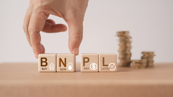 Hand arranged wooden cube blocks with  BNPL, alphabets abbreviation , and icon with blurred stack of  coins. For buy now pay later online shopping concept