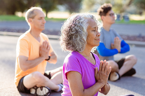 An active and healthy senior woman of Pacific Islander ethnicity sits with her Caucasian husband on the ground of a park as they practice meditation during a group yoga class on a beautiful day.