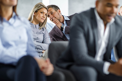 Young businesswoman listening to her colleague telling her a secret on a seminar in board room. Focus is on man.