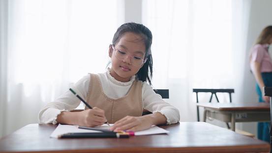 Asian school girl in glasses studying, writing notes in notebook at classroom. Schoolgirl studying and preparing for exams. little pupil writing at desk in classroom. Education knowledge concept