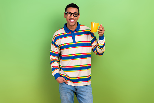 Photo portrait of handsome young guy holding yellow tea cup drinking dressed stylish striped outfit isolated on green color background.